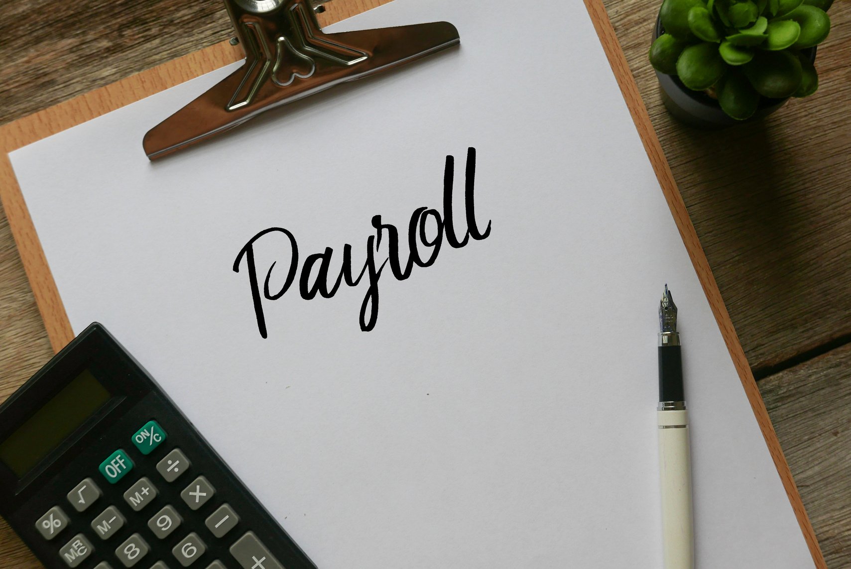 Top view of plant,calculator,pen and clipboard with white paper written with Payroll on wooden background.